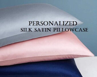 May include: Three silk satin pillowcases in shades of grey, pink, and navy blue are stacked. The top pillowcase is grey, the middle one is pink, and the bottom one is navy. Text on the image reads: PERSONALIZED SILK SATIN PILLOWCASE.