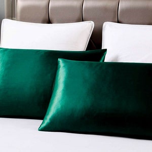 May include: Two emerald green satin pillowcases on a white bed. The pillowcases have a lustrous sheen and are rectangular. Two white pillows are in the background, along with a neutral-toned headboard.