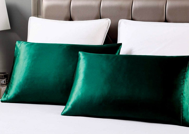 May include: Two emerald green satin pillowcases on a white bed. The pillowcases have a lustrous sheen and are rectangular. Two white pillows are in the background, along with a neutral-toned headboard.