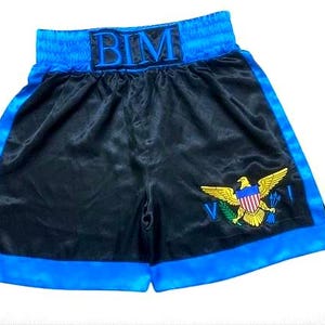 Op de afbeelding: Zwarte boksshorts met blauwe randen en tailleband. De tailleband heeft de letters "BIM" in blauw. Een geborduurd embleem van een gouden adelaar met een schild en olijftak staat op de short.