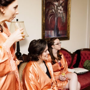 May include: Three women in peach-colored satin robes are gathered indoors. One is drinking from a straw, while the others sit on a red velvet sofa. A painting hangs on the wall behind them, and a lamp is visible.