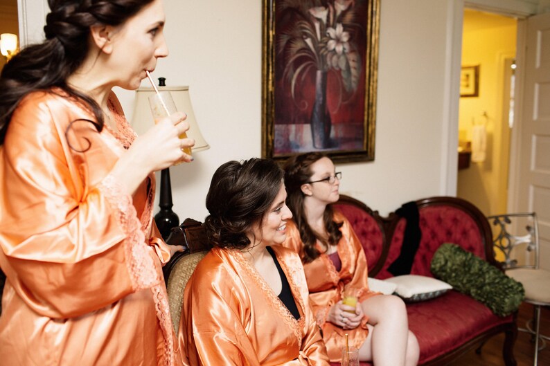 May include: Three women in peach-colored satin robes are gathered indoors. One is drinking from a straw, while the others sit on a red velvet sofa. A painting hangs on the wall behind them, and a lamp is visible.