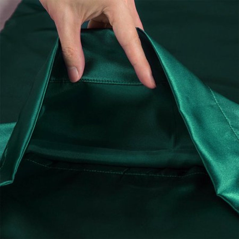 May include: Close-up of a deep emerald green satin fabric. The fabric is smooth and shiny, with a slight sheen. A hand is gently touching the fabric, highlighting its texture and drape. The image focuses on the luxurious quality of the material.