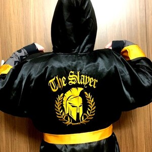 以下が含まれることがあります： 背面に月桂樹の冠に囲まれた黄色のスパルタヘルメットの刺繍エンブレムがあしらわれた、黒いサテン地のフード付きローブ。エンブレムの上には「The Slayer」という文字が黄色で刺繍されています。ローブには黄色のウエストベルトが付いています。