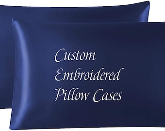 May include: Two navy blue satin pillowcases. The pillowcases have the words "Custom Embroidered Pillow Cases" embroidered in white. The pillowcases are rectangular and have a smooth, shiny texture.