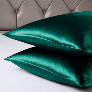 May include: Two emerald green satin pillowcases are stacked on a white surface. The pillowcases have a lustrous sheen, reflecting light. The background features a grey, tufted headboard. The pillowcases are a luxurious bedding item.