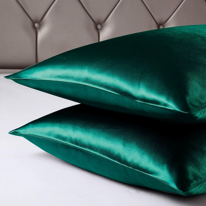 May include: Two emerald green satin pillowcases are stacked on a white surface. The pillowcases have a lustrous sheen, reflecting light. The background features a grey, tufted headboard. The pillowcases are a luxurious bedding item.