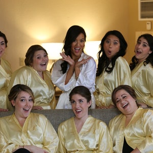 May include: A group of women wearing satin robes in shades of yellow and white, posing for a photo. The robes have a monogram on the front. The women are smiling and laughing.