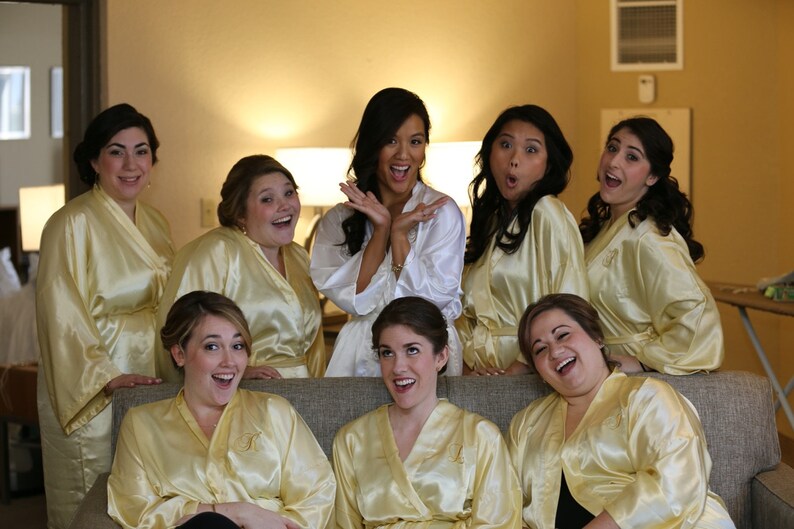 May include: A group of women wearing satin robes in shades of yellow and white, posing for a photo. The robes have a monogram on the front. The women are smiling and laughing.