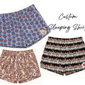 Puede incluir: Tres pares de pantalones cortos de dormir personalizados. Un par es azul claro con un estampado floral y un parche de oso de peluche. Otro par es marrón con un diseño de cachemira. El tercer par es negro con un patrón de figuras repetidas y un parche de corazón.