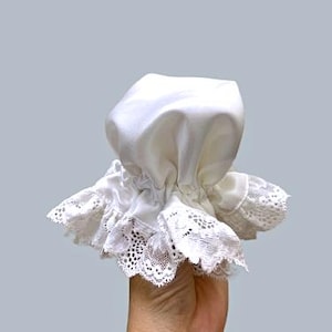 Pode incluir: Um gorro de cetim branco com uma borda de renda com babados. O gorro é franzido na parte superior e segurado em uma mão contra um fundo cinza. A borda de renda tem uma borda delicada e ondulada.