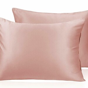 May include: Two blush-pink satin pillowcases. The rectangular pillows have a smooth, shiny finish. The pillowcases are displayed on a white background. The pillowcases are a solid colour and appear to be made of a soft material.