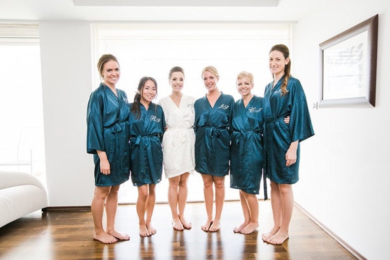 May include: A group of six people wearing robes. Five are in teal robes with personalized initials embroidered on the chest. One person is in a white robe. The group is standing on a wooden floor in a well-lit room.
