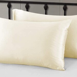 May include: Two ivory silk pillowcases on a white bed. The rectangular pillowcases have a smooth, shiny texture. A black metal headboard and white bed sheet are in the background.