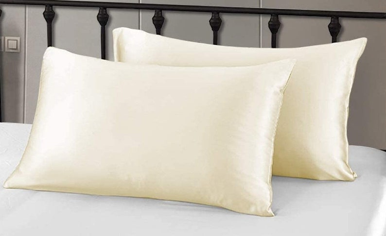May include: Two ivory silk pillowcases on a white bed. The rectangular pillowcases have a smooth, shiny texture. A black metal headboard and white bed sheet are in the background.