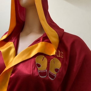 May include: A burgundy satin boxing robe with a gold satin trim and hood. The robe features embroidered gold boxing gloves and the name "US" in gold thread. The robe is designed with a wrap-around style.