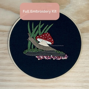 May include: A full embroidery kit featuring a whimsical design of a snail, mushroom, and grass on a dark blue background. The embroidery is set in a wooden hoop. The text "Full Embroidery Kit" is displayed at the top.