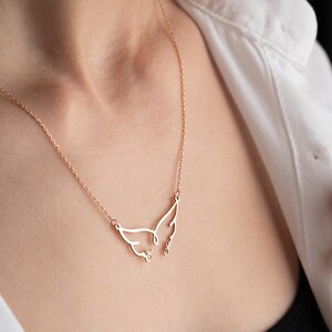 May include: A delicate rose gold necklace featuring a stylized cat face pendant. The pendant has a minimalist design with wing-like shapes and small details. The necklace is worn against a white shirt.