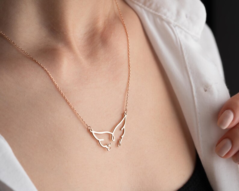 May include: A delicate rose gold necklace featuring a stylized cat face pendant. The pendant has a minimalist design with wing-like shapes and small details. The necklace is worn against a white shirt.