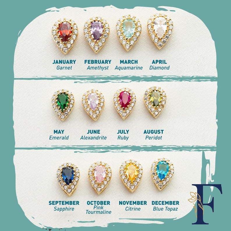 May include: A collection of twelve teardrop-shaped birthstone pendants. Each pendant features a different gemstone color, including red garnet, purple amethyst, and blue sapphire, set in a gold-colored frame with surrounding clear crystals. The months of the year are labeled below each pendant.