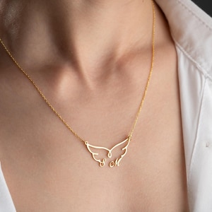 May include: A gold-colored necklace featuring a delicate wing design. The pendant includes the initials "S M" in a cursive font. The necklace is worn against a white shirt.