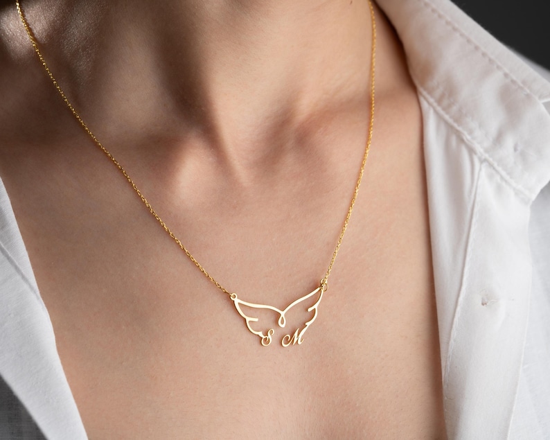 May include: A gold-colored necklace featuring a delicate wing design. The pendant includes the initials "S M" in a cursive font. The necklace is worn against a white shirt.