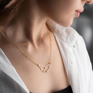 May include: A delicate gold-colored necklace featuring a wing-shaped pendant. The pendant has an open design with two stylized wings and two small letters. The necklace is worn against a white shirt.