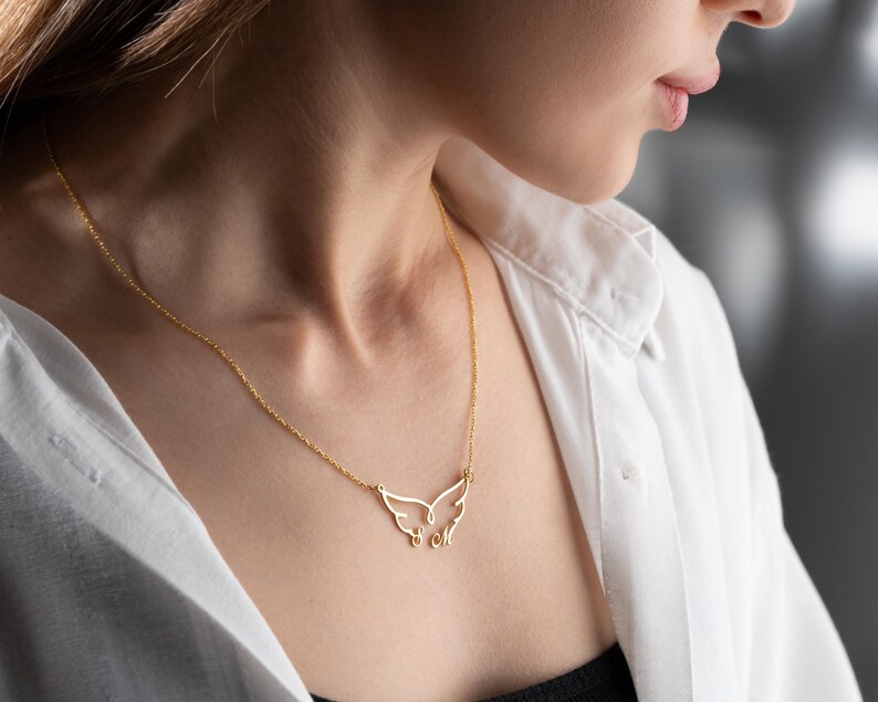 May include: A delicate gold-colored necklace featuring a wing-shaped pendant. The pendant has an open design with two stylized wings and two small letters. The necklace is worn against a white shirt.