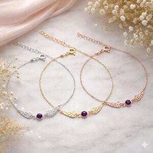 May include: Three delicate bracelets in silver, gold, and rose gold. Each features a wing design and a small, round, purple gemstone. The bracelets are displayed on a marble surface with soft lighting and floral accents.