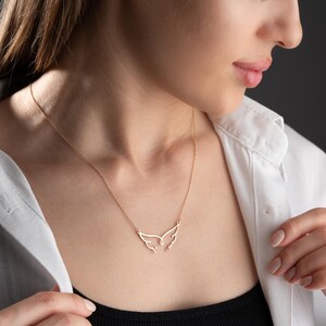 May include: A delicate gold-colored necklace featuring a wing-shaped pendant. The necklace is worn against a white collared shirt and a black top. The pendant's design is minimalist, with the wings spread open.