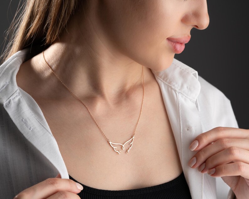 May include: A delicate gold-colored necklace featuring a wing-shaped pendant. The necklace is worn against a white collared shirt and a black top. The pendant's design is minimalist, with the wings spread open.