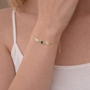 May include: A gold-colored bracelet featuring a central, round, dark green gemstone. The bracelet has delicate wing-shaped designs on either side of the gemstone. The bracelet is on a person's wrist.