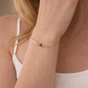 May include: A gold-colored bracelet featuring a central red gemstone flanked by delicate wing designs. The bracelet is attached to a fine chain, creating an elegant and refined accessory.