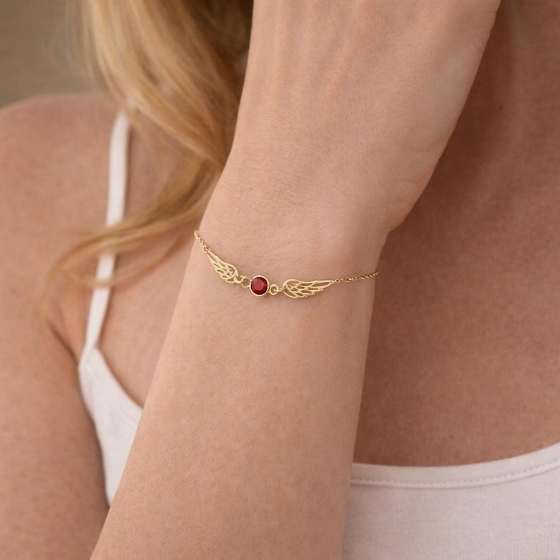 May include: A gold-colored bracelet featuring a central red gemstone flanked by delicate wing designs. The bracelet is attached to a fine chain, creating an elegant and refined accessory.