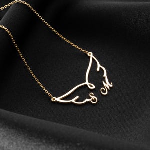 May include: A delicate gold-colored necklace featuring a wing design with the initials "S" and "M". The necklace is displayed on a black fabric background, highlighting the intricate details of the pendant and chain.