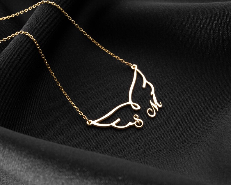May include: A delicate gold-colored necklace featuring a wing design with the initials "S" and "M". The necklace is displayed on a black fabric background, highlighting the intricate details of the pendant and chain.