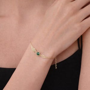 May include: A gold-colored bracelet featuring a central green gemstone and delicate wing-shaped accents. The bracelet is attached to a fine chain, creating an elegant and refined look. The bracelet is worn on a wrist.