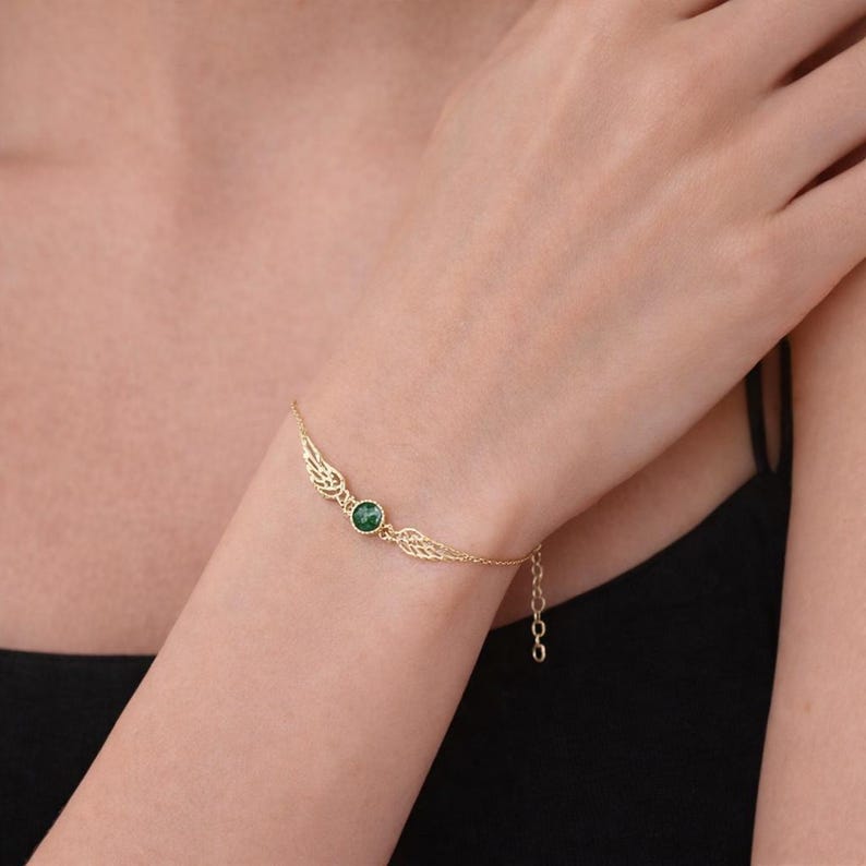 May include: A gold-colored bracelet featuring a central green gemstone and delicate wing-shaped accents. The bracelet is attached to a fine chain, creating an elegant and refined look. The bracelet is worn on a wrist.