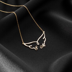 May include: A gold-colored necklace with a wing design and the letters "S M". The necklace is displayed on a black, textured fabric. The chain is delicate and the pendant is a stylized wing shape.