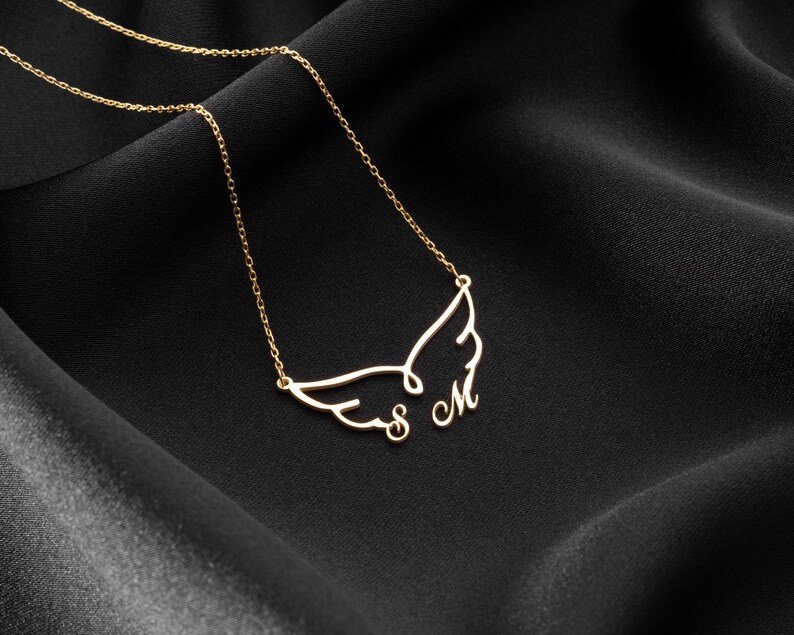 May include: A gold-colored necklace with a wing design and the letters "S M". The necklace is displayed on a black, textured fabric. The chain is delicate and the pendant is a stylized wing shape.