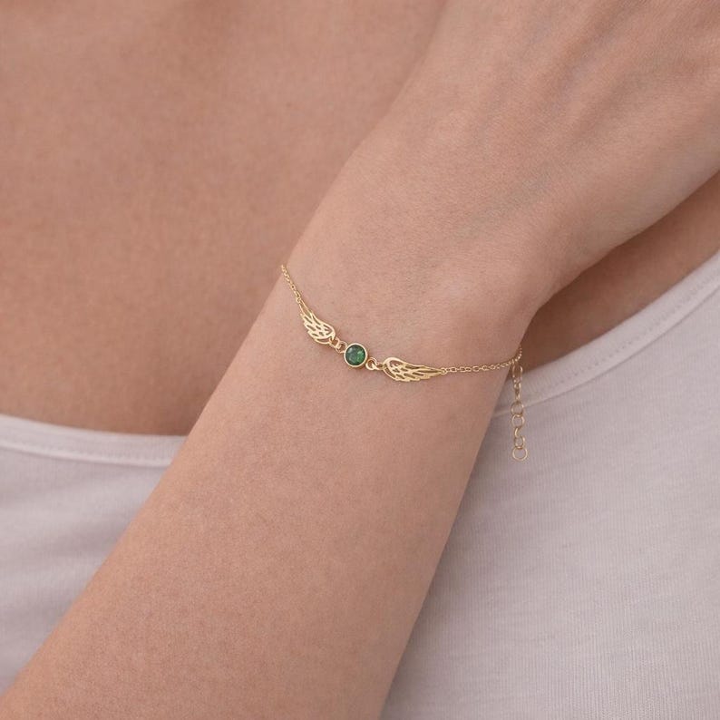 May include: A gold-colored bracelet featuring a central green gemstone and delicate wing-shaped designs on either side. The bracelet is shown on a wrist, highlighting its fine details and elegant aesthetic. The adjustable chain adds to its versatility.