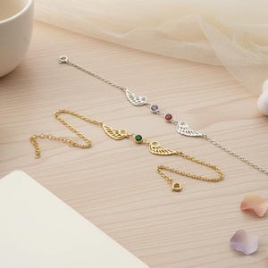 May include: Two delicate bracelets with wing designs. One is gold-toned with a green gemstone, the other is silver-toned with purple and red gemstones. The bracelets are displayed on a light wood surface with a cup and saucer.