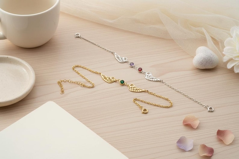 May include: Two delicate bracelets with wing designs. One is gold-toned with a green gemstone, the other is silver-toned with purple and red gemstones. The bracelets are displayed on a light wood surface with a cup and saucer.