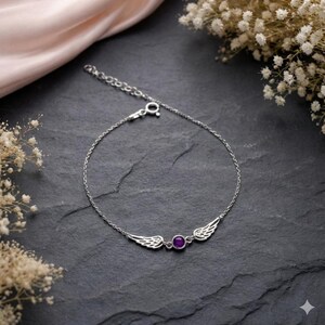 May include: A silver bracelet featuring a pair of detailed wings flanking a round, purple gemstone. The bracelet is on a dark, textured surface, with a soft pink fabric and white flowers in the background.