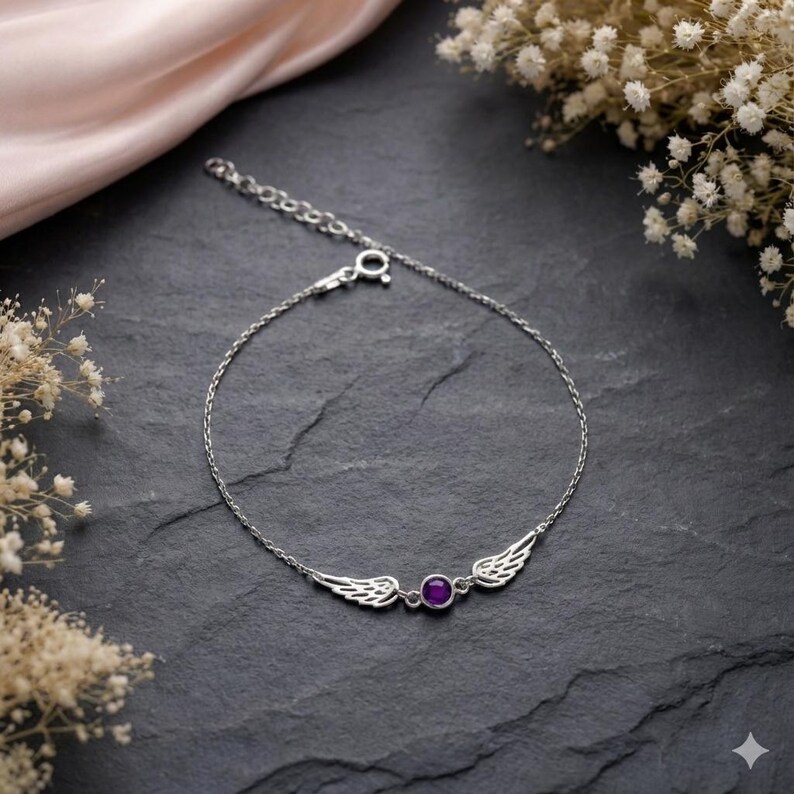 May include: A silver bracelet featuring a pair of detailed wings flanking a round, purple gemstone. The bracelet is on a dark, textured surface, with a soft pink fabric and white flowers in the background.