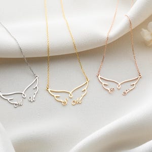 May include: Three delicate necklaces with wing-shaped pendants, each with personalized initials. The necklaces are in silver, gold, and rose gold. The pendants are suspended from fine chains, resting on a white fabric background.