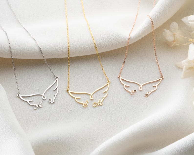 May include: Three delicate necklaces with wing-shaped pendants, each with personalized initials. The necklaces are in silver, gold, and rose gold. The pendants are suspended from fine chains, resting on a white fabric background.