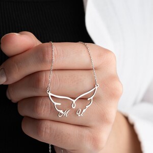 May include: A silver-colored necklace featuring a wing-shaped pendant with the letters "M" and "U" engraved. The necklace is held by a person, showcasing the delicate chain and the personalized design. The pendant has a polished finish.