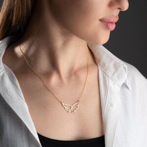 May include: A delicate gold-colored necklace featuring a wing-shaped pendant. The pendant has intricate cut-out details, and the necklace is worn against a white collared shirt and a black top.
