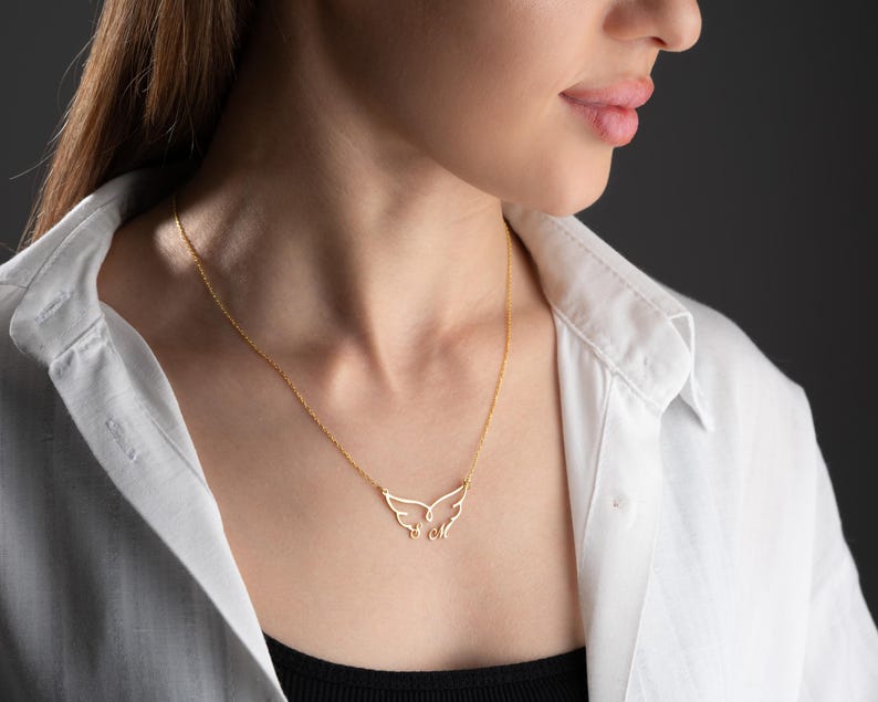 May include: A delicate gold-colored necklace featuring a wing-shaped pendant. The pendant has intricate cut-out details, and the necklace is worn against a white collared shirt and a black top.